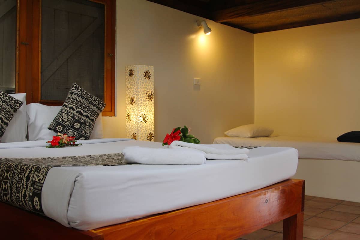 A hotel room with a wooden bed, two white beds, a decorative wall lamp, and a window with a wooden frame.
