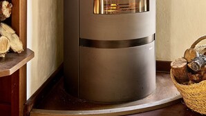 A modern wood-burning stove with a glass door, a black chimney, and a wooden shelf with a bottle and a teddy bear.