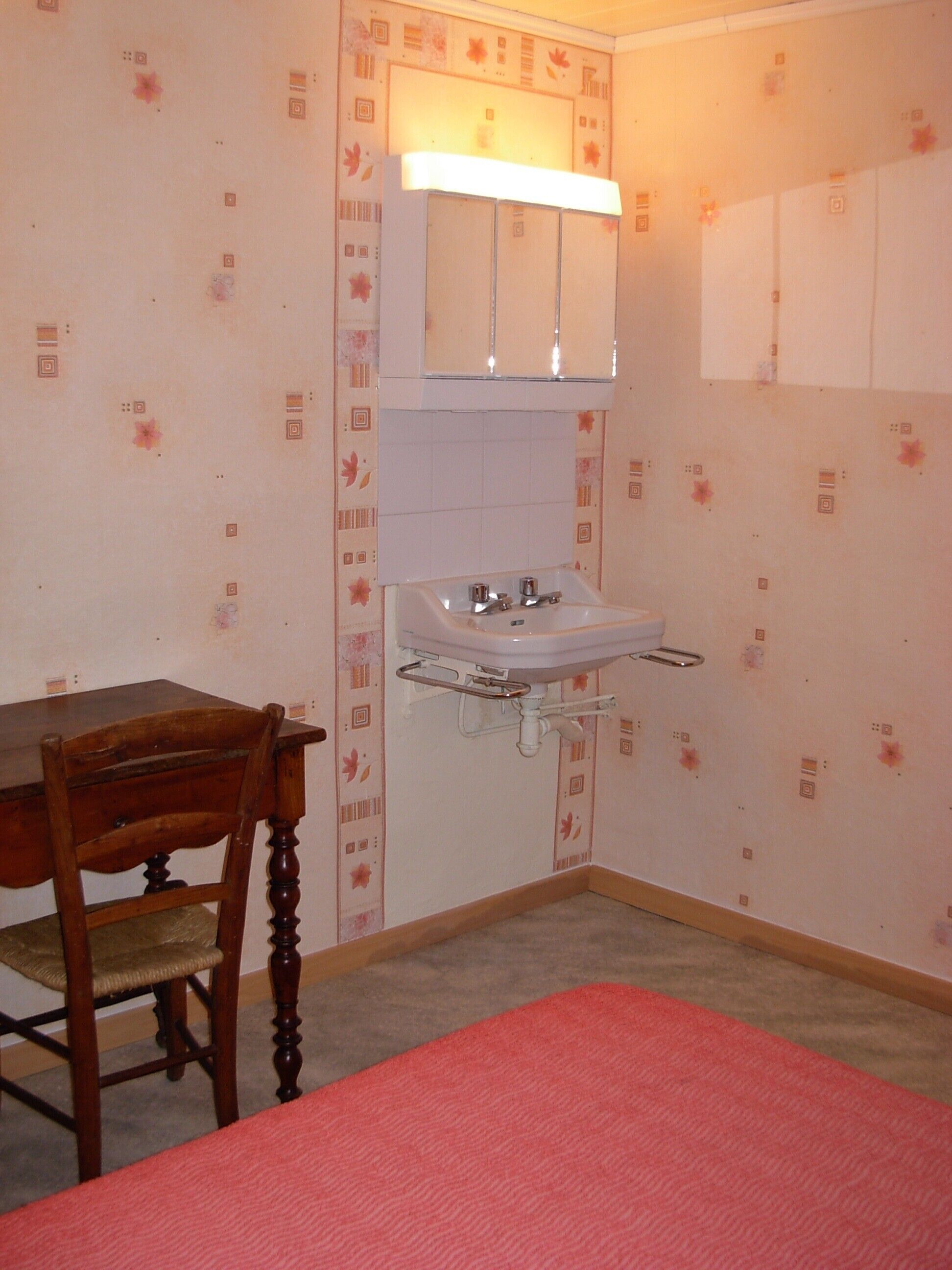 Ein kleines Badezimmer mit Waschbecken, Spiegel und Schrank. Ein Holzstuhl und ein Tisch mit roter Tischdecke stehen dort.