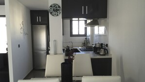 Una cocina moderna con armarios oscuros, un refrigerador de acero inoxidable, una máquina de café y un comedor con sillas blancas.