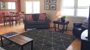 A living room with a dark grey sofa, a wooden coffee table, and a black rug. There is a dining table with chairs, a ceiling fan, and framed pictures on the wall. - Waterfront Paradise with  NEW Jacuzzi Spa! (Canandaigua)