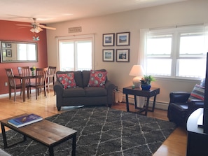 A living room with a dark grey sofa, a wooden coffee table, and a black rug. There is a dining table with chairs, a ceiling fan, and framed pictures on the wall. - Waterfront Paradise with  NEW Jacuzzi Spa! (Canandaigua)