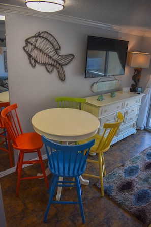 A dining area with a round table and colorful chairs, a fish wall art, a flat-screen TV, and a white cabinet with drawers.