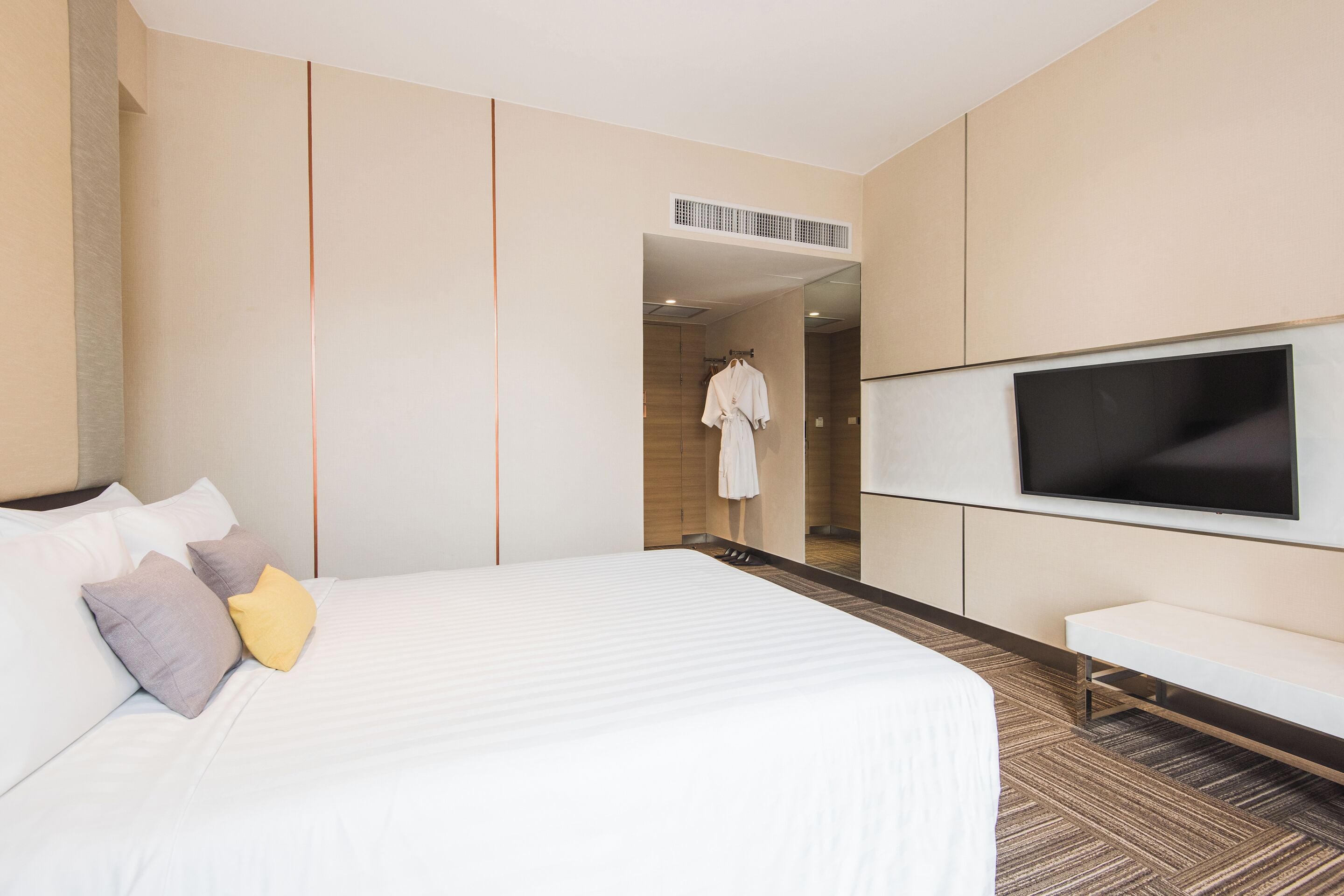 A hotel room with a large bed, a flat-screen TV mounted on the wall, a bench, and a robe hanging on a rack.
