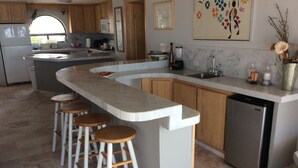 Uma cozinha moderna com uma ilha sinuosa, banquetas de madeira, uma máquina de lavar louça e uma janela com vista.