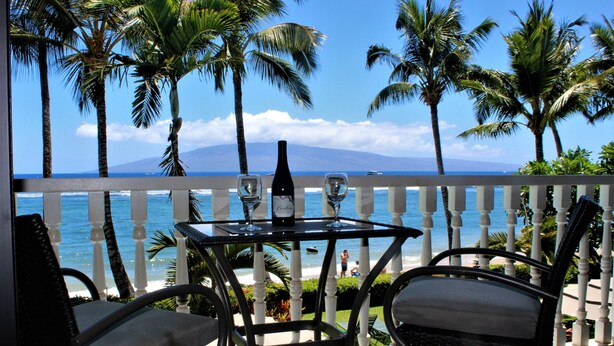 Un balcon avec une table et des chaises, donnant sur une plage bordée de palmiers et sur l’océan.