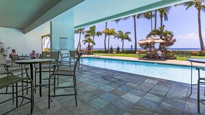 Un espace aménagé au bord de la piscine, avec des tables et des chaises, un panneau indiquant les règles de la piscine, et une vue sur l’océan bordée de palmiers.