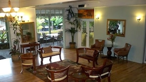 Un salon spacieux doté d’un plancher en bois, d’une table basse centrale et de plusieurs chaises en bois. On aperçoit un coin repas à travers une porte ouverte.