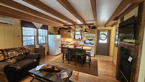 A cozy living room with a sofa, coffee table, and fireplace. A kitchen with wooden cabinets and a dining area is visible in the background. - The Ole Bear Rustic Mountain Cabin (Tellico Plains)
