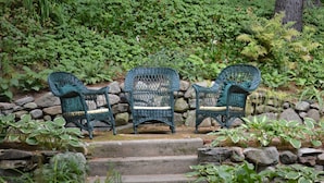 Drei Korbstühle mit Kissen an einer Steinsäule, umgeben von üppigem Grün und Stufen, die zu einem Garten hinaufführen.