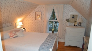 Ein Schlafzimmer mit einem Bett, einem Schminktisch, einem Nachttisch, einem Fenster mit Vorhängen und einem gerahmten Bild an der Wand.