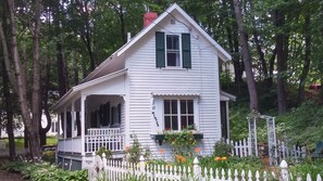 Une maison en bois blanche, avec des volets verts, un porche et une clôture en bois peinte en blanc.