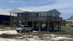 Uma casa de praia de dois andares com varanda, um carro estacionado na frente e uma praia de areia ao fundo.