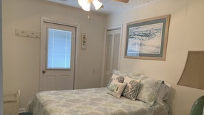 Quarto com cama, ventilador de teto, janela com persianas, porta e um quadro na parede.