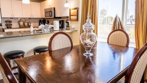 A dining area with a wooden table, four chairs, a glass jar, and a kitchen in the background. - BEACHFRONT GRAND PANAMA, 1st FLR DIRECT TO BEACH, PET-FRIENDLY. NEXT TO TIKI BAR (Panama City Beach)