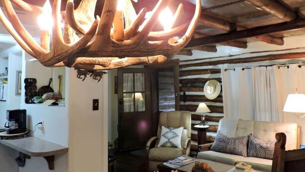 A rustic interior with a large antler chandelier, wooden beams, a cozy seating area, and a kitchen with a coffee machine. - Cabin (Prescott)