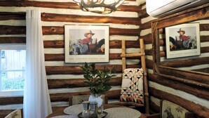 A rustic cabin interior with a wooden log wall, a chandelier, framed artwork, a dining table with placemats, and a window with curtains. - Cabin (Prescott)