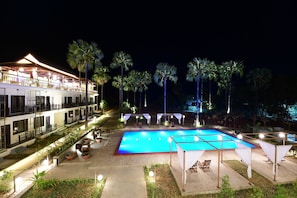 Una piscina con sillas de descanso y sombrillas alrededor, un edificio con balcones y palmeras.