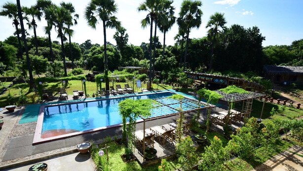 Una piscina rodeada de exuberante vegetación y palmeras, con sillones de descanso y un pabellón.