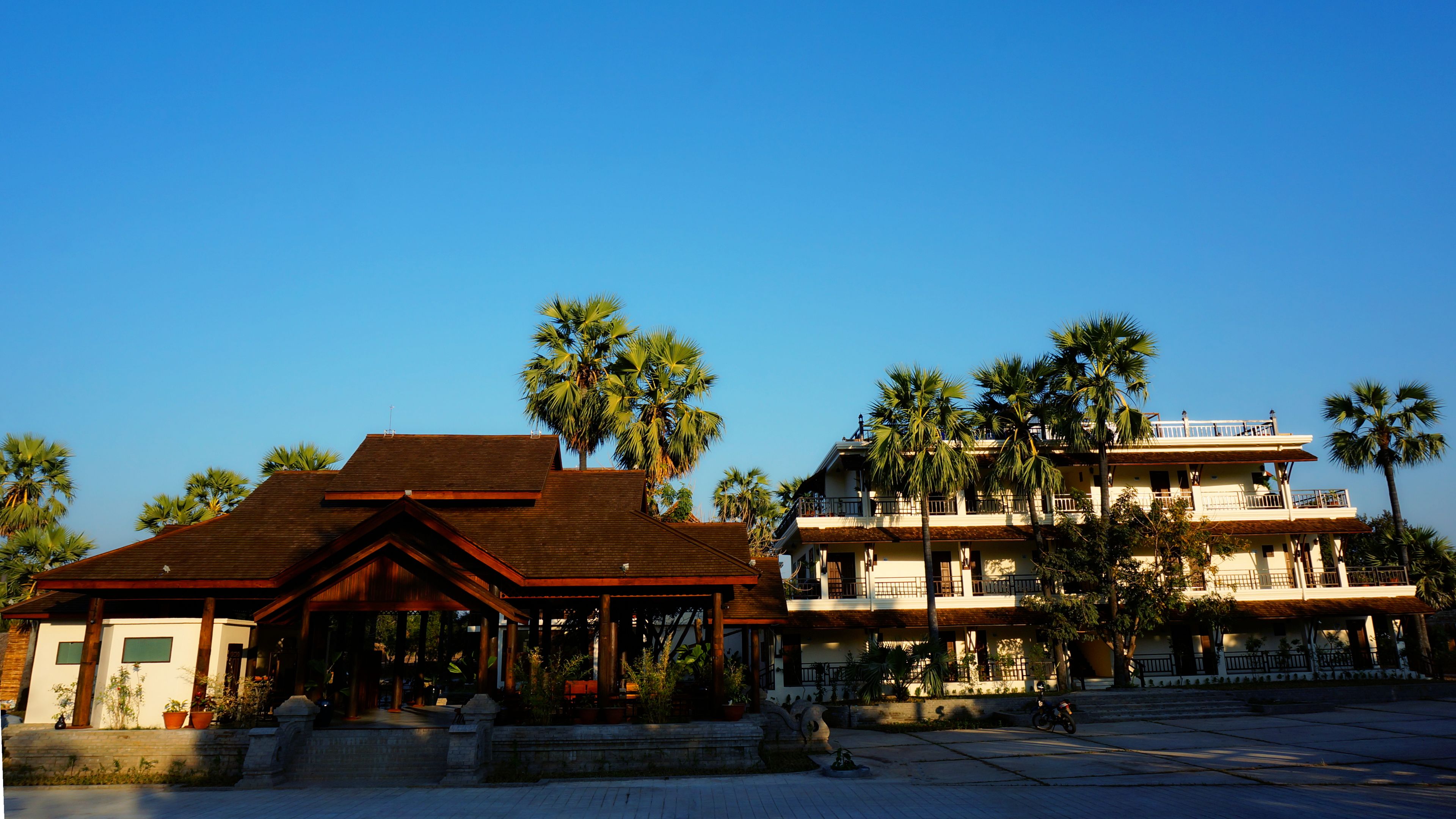 A two-story building with a wooden roof and multiple balconies, surrounded by palm trees.