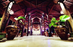 Um hall amplo com teto alto e vigas de madeira, destacando dois grandes vasos com a inscrição 'Myanmar Hotel'.