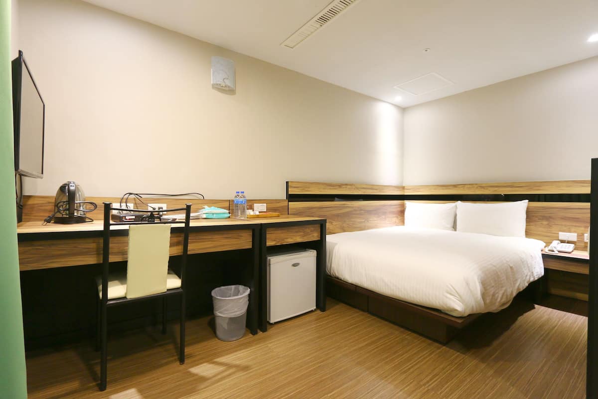 Hotellihuone, jossa on sänky, työpöytä tuolilla, pieni jääkaappi ja roskakori.