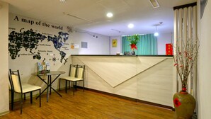 Zona de recepción con un mapa mundial en la pared, mostrador de recepción, dos sillones, una mesa y un jarrón con ramas.