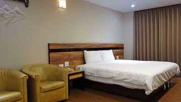 A hotel room with a wooden headboard bed, a bedside table with a phone, a chair, and a wall-mounted light fixture.
