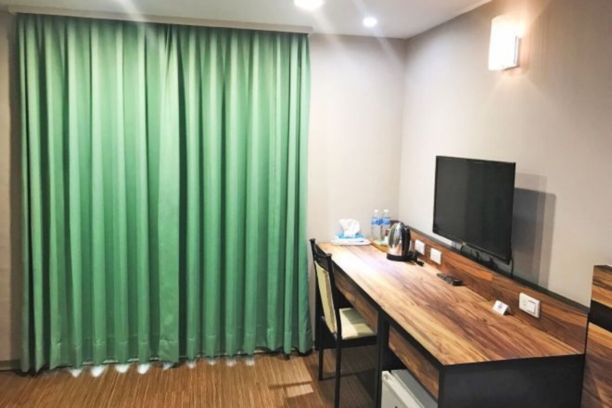 A room with a wooden desk, a flat-screen TV, a green curtain, and a wall-mounted light.