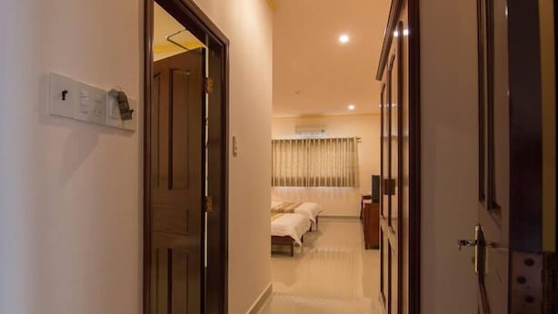 A hotel corridor with a wooden door on the left, a bed with a white bedspread in the background, and a small table with a lamp. - Nhat Ha Hotel (Can Tho)