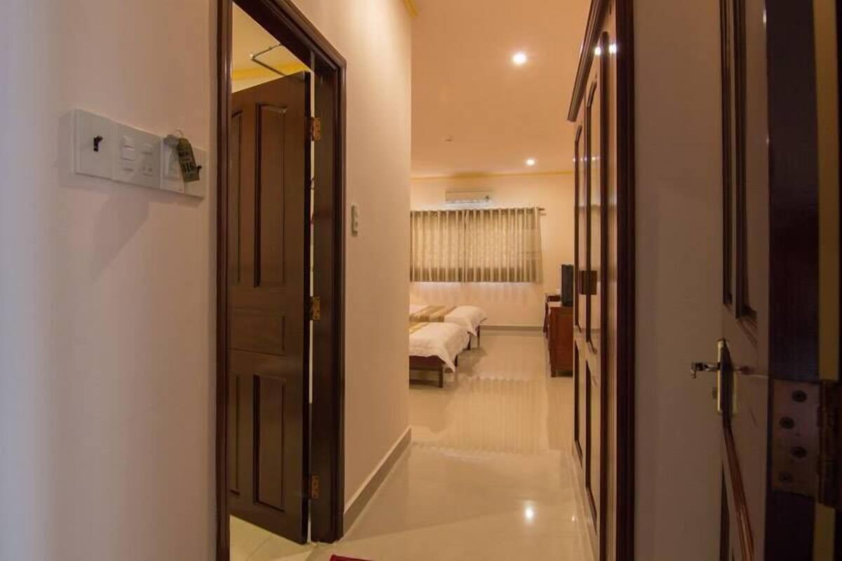 Un pasillo de hotel con una puerta de madera a la izquierda, una cama con colcha blanca al fondo y una mesita con una lámpara.