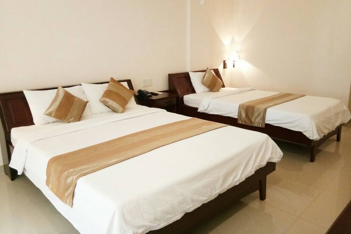 Habitación de hotel con dos camas individuales, cada una con cabecero de madera, ropa de cama blanca y una manta marrón. Hay una mesita de noche con una lámpara entre las camas.