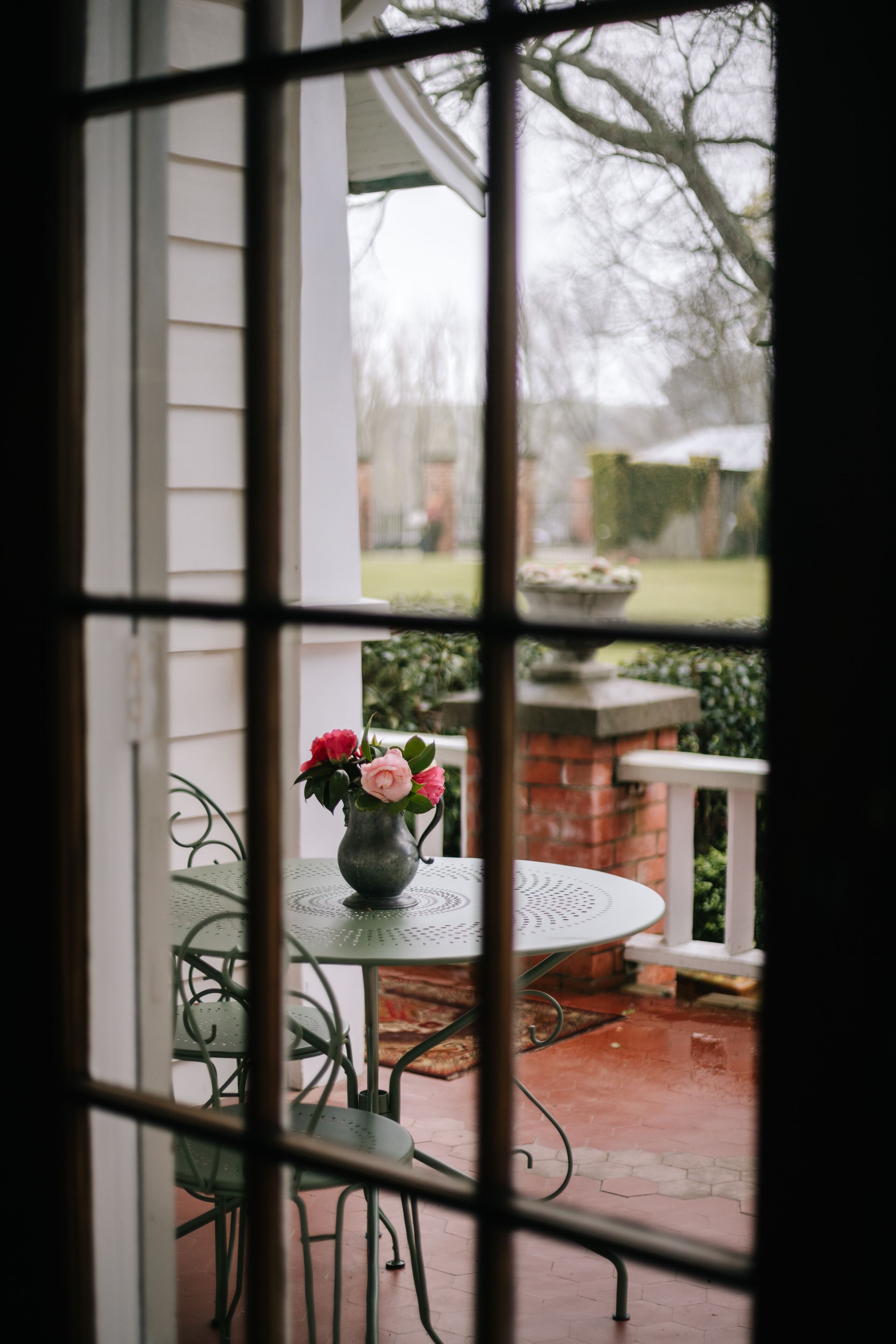 A view through a window with a metal grill, featuring a round table with a vase of flowers, green chairs, and a brick structure in the background.