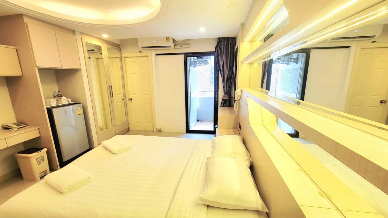 Una habitación de hotel con una cama, un escritorio con computadora, un basurero y una ventana con cortinas.