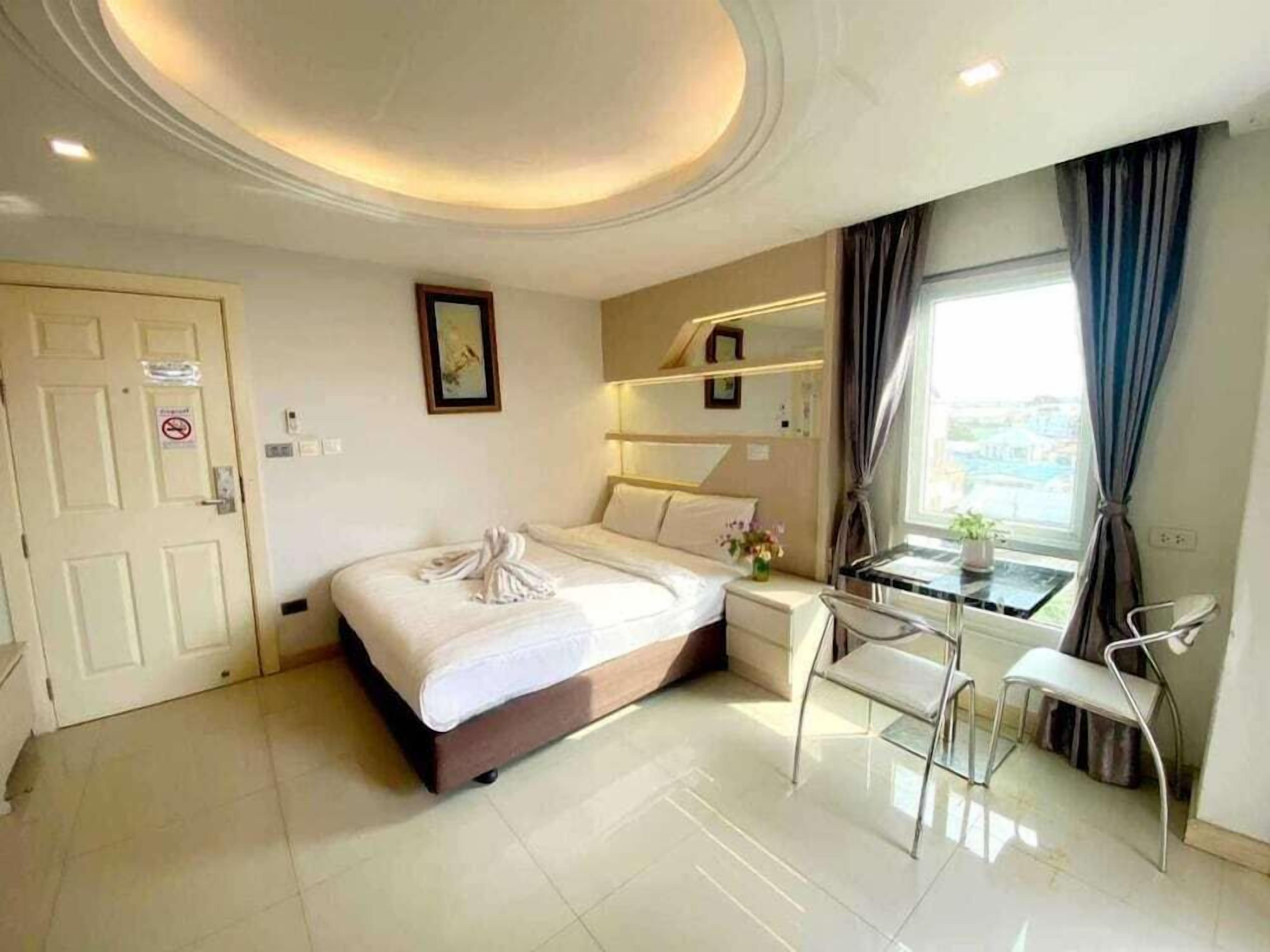 A hotel room with a bed, a desk, a chair, a window with curtains, a door, and a ceiling with recessed lighting.