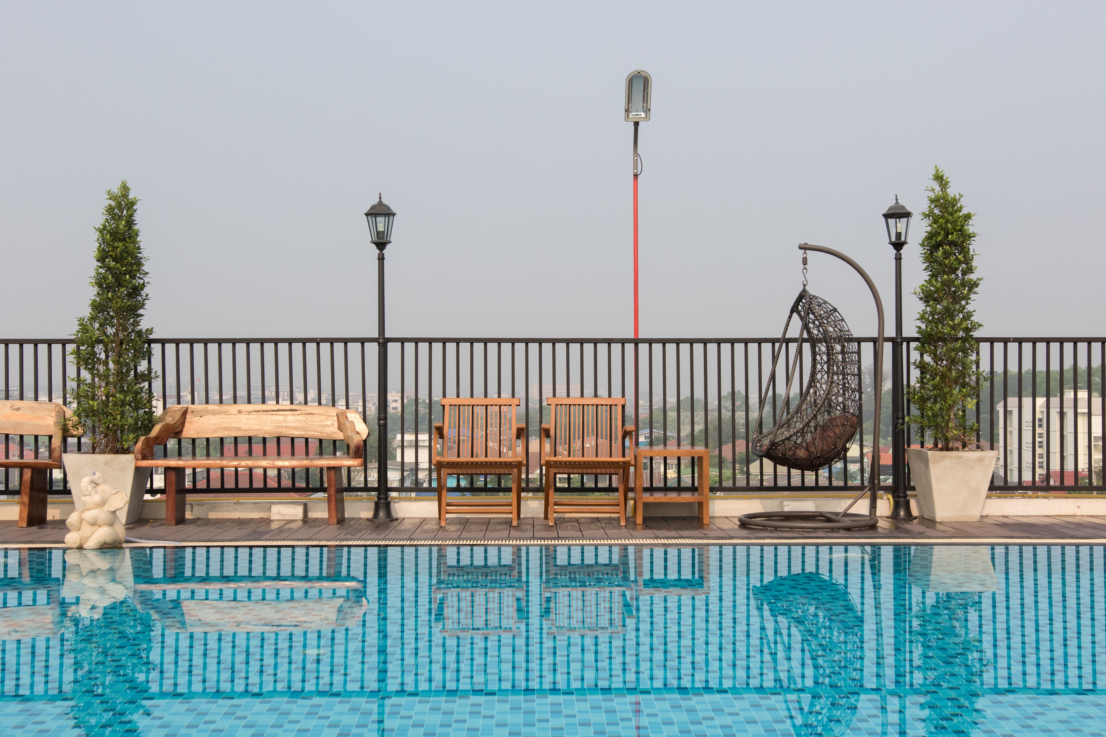 A rooftop pool area with wooden chairs, a hanging chair, and potted plants.