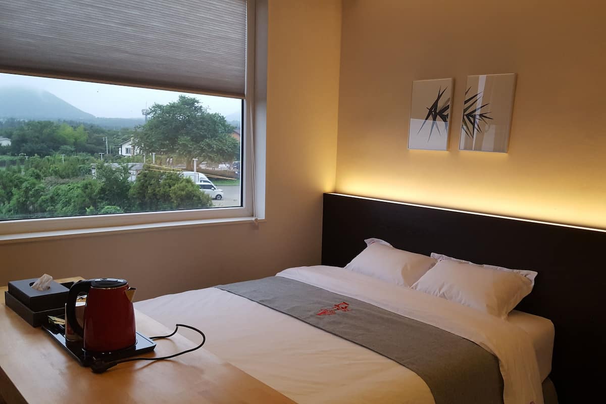 A hotel room with a bed, a coffee maker, a window with a view of greenery and a mountain, and two wall art pieces.