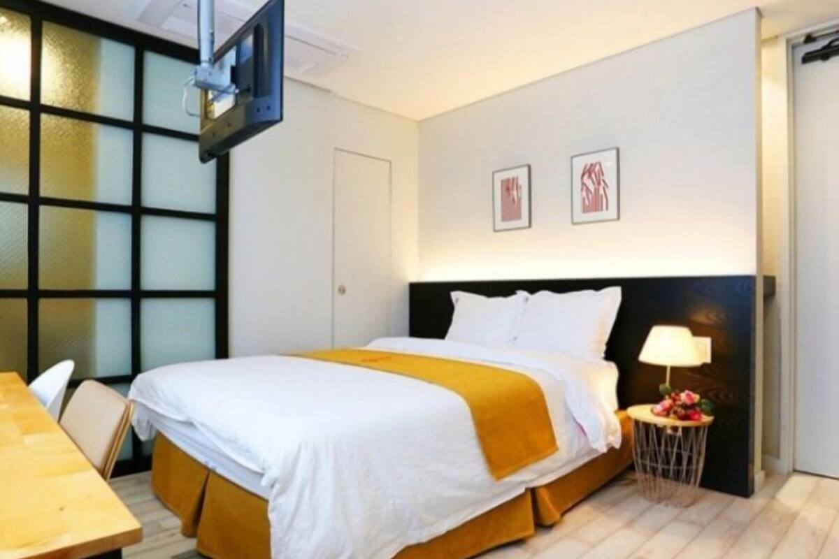 A hotel room with a bed, a desk, a chair, a TV mounted on the wall, and a small table with a lamp and flowers.