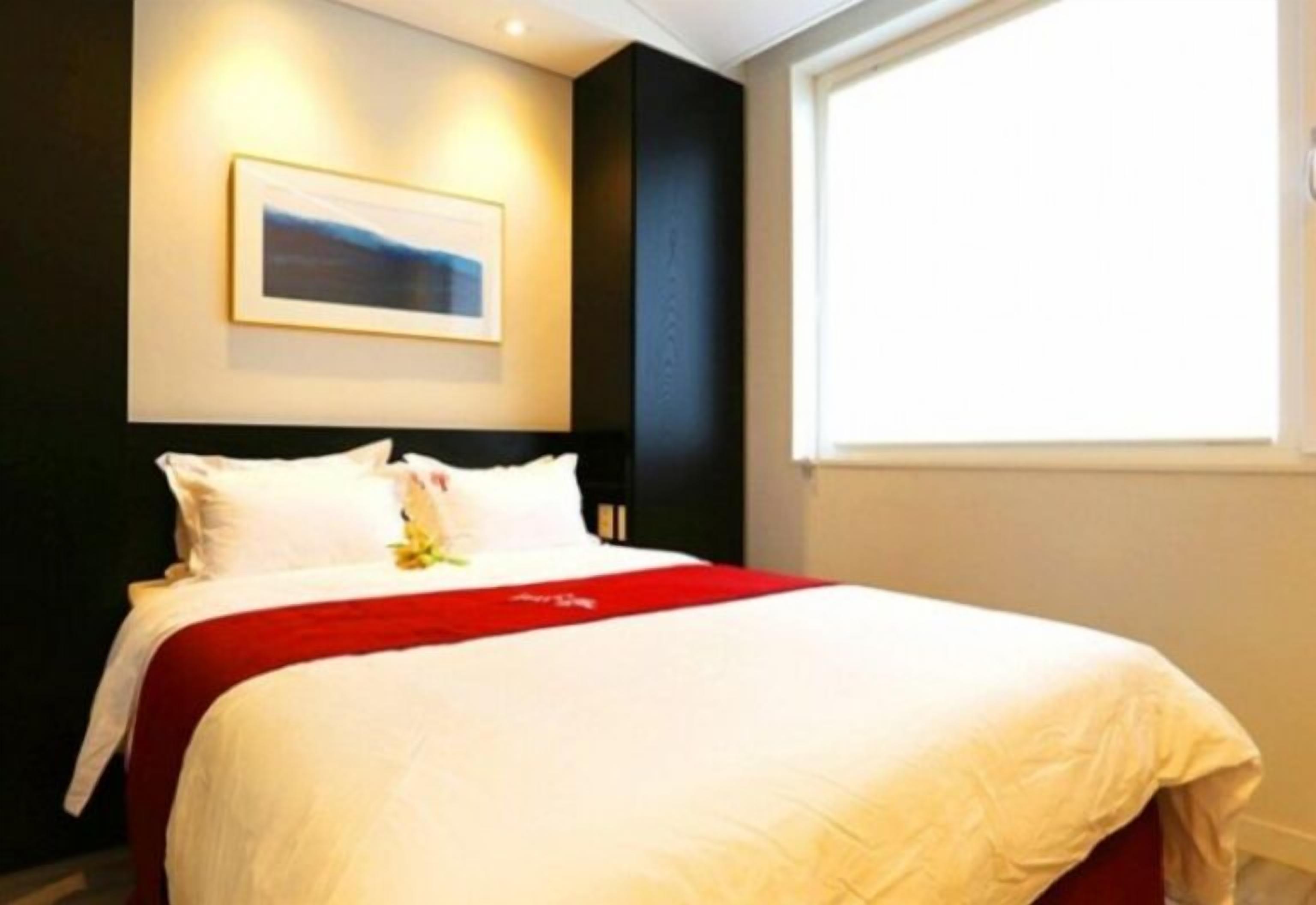 Ein ordentlich bezogenes Bett mit weißer und roter Bettwäsche, ein gerahmtes Bild an der Wand und ein Fenster, das für natürliches Licht sorgt.