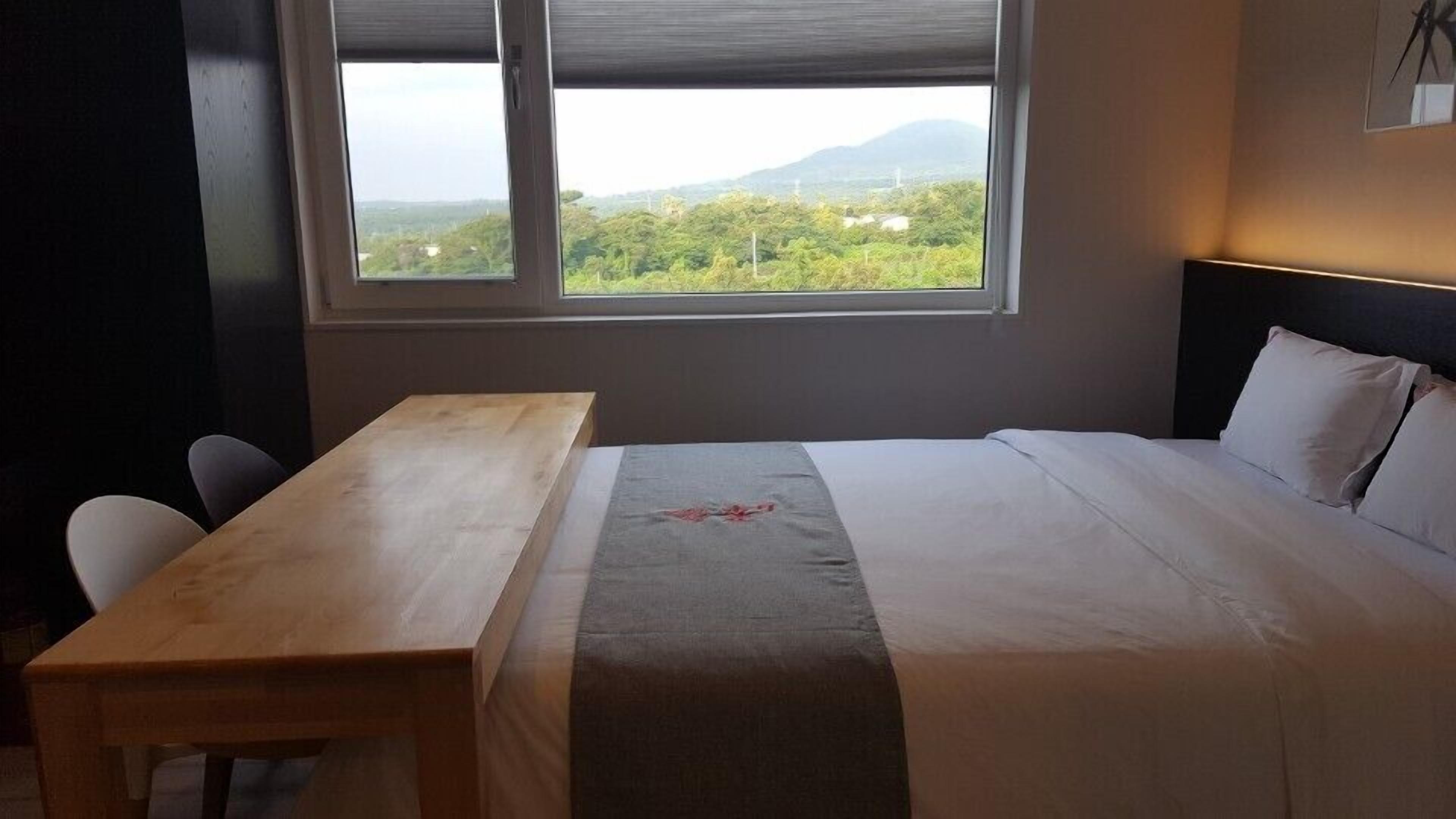 A hotel room with a bed, a wooden table, and a window offering a view of greenery and mountains.