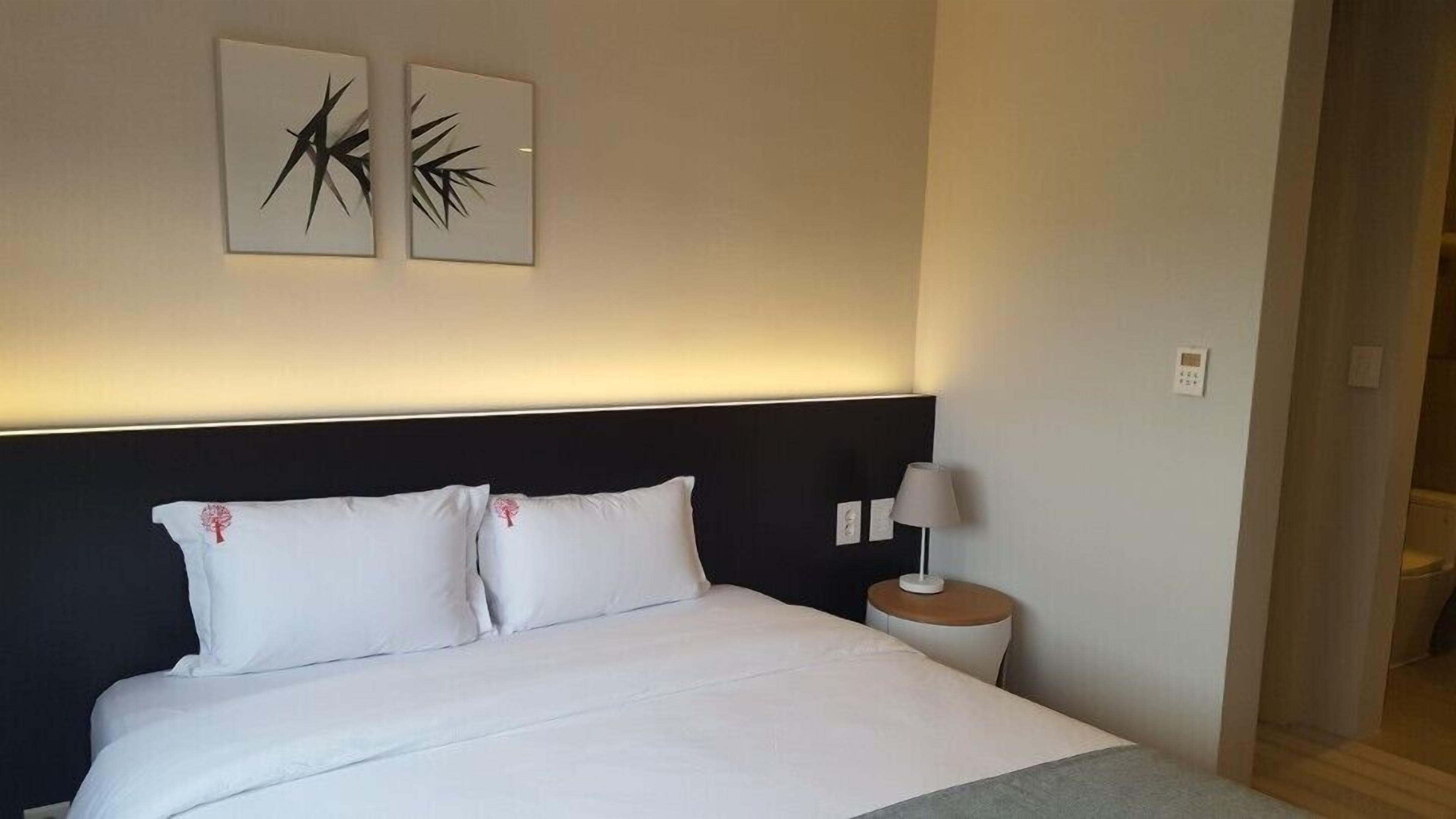 Una cama bien tendida con sábanas blancas y dos almohadas, una mesita de noche con lámpara y dos obras de arte en la pared con motivos abstractos de plantas.