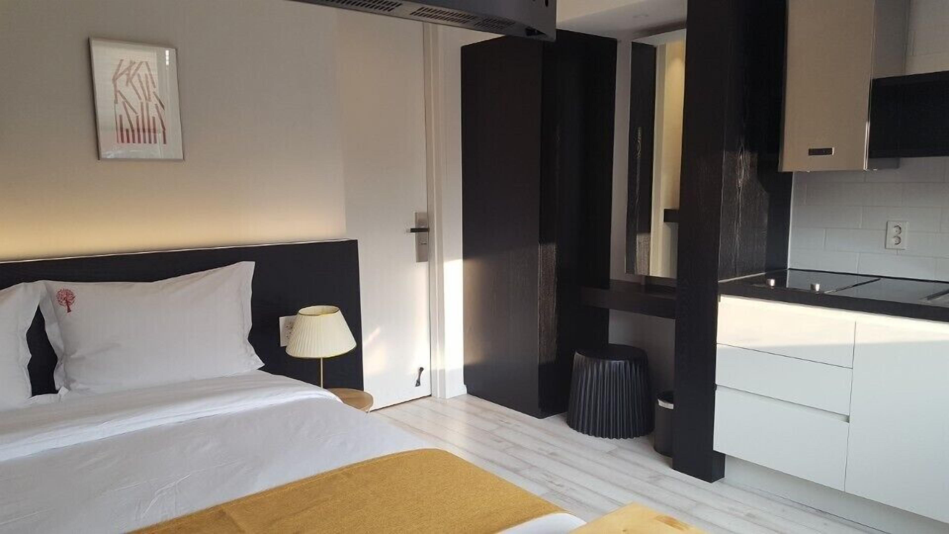 A modern hotel room with a bed, bedside table, lamp, and a view of the kitchen.