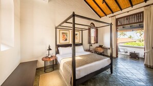 Uma cama com dossel e estrutura de madeira, uma mesa redonda pequena com uma luminária, e vista para o verde lá fora.
