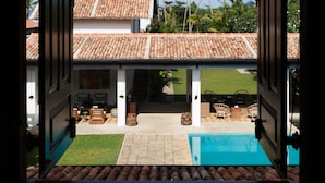 Uma porta aberta conduzindo a uma casa com telhado de cerâmica, piscina e palmeiras ao fundo.