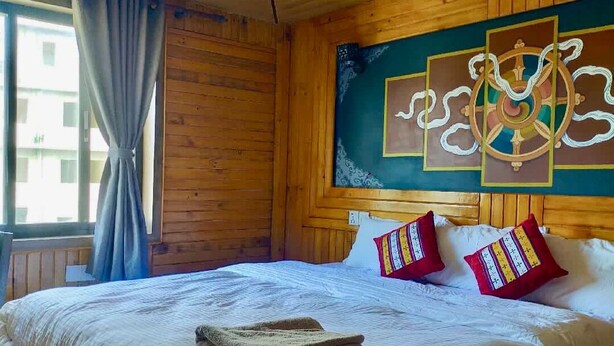 A wooden room with a bed, a window with curtains, and a wall with a decorative painting. - Hotel Grand Shambala (Muktinath)