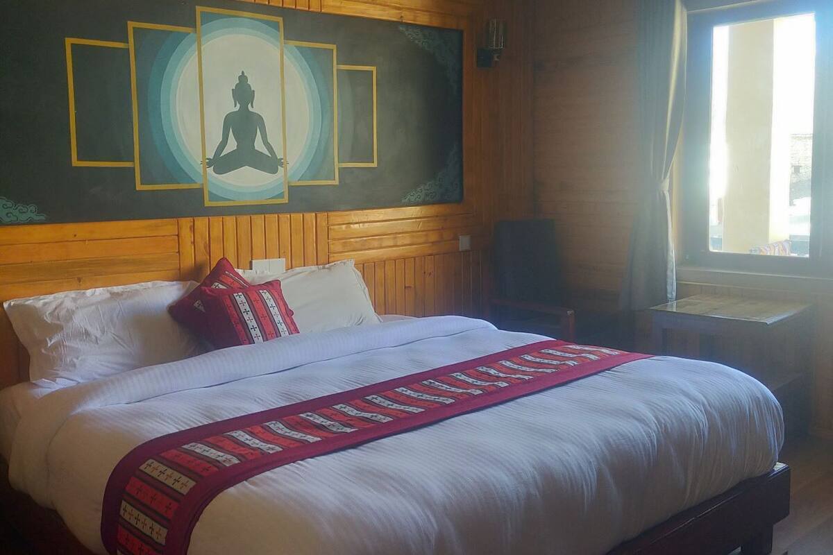 A room with a large bed, wooden walls, a window with curtains, and a wall-mounted artwork of a meditating figure.