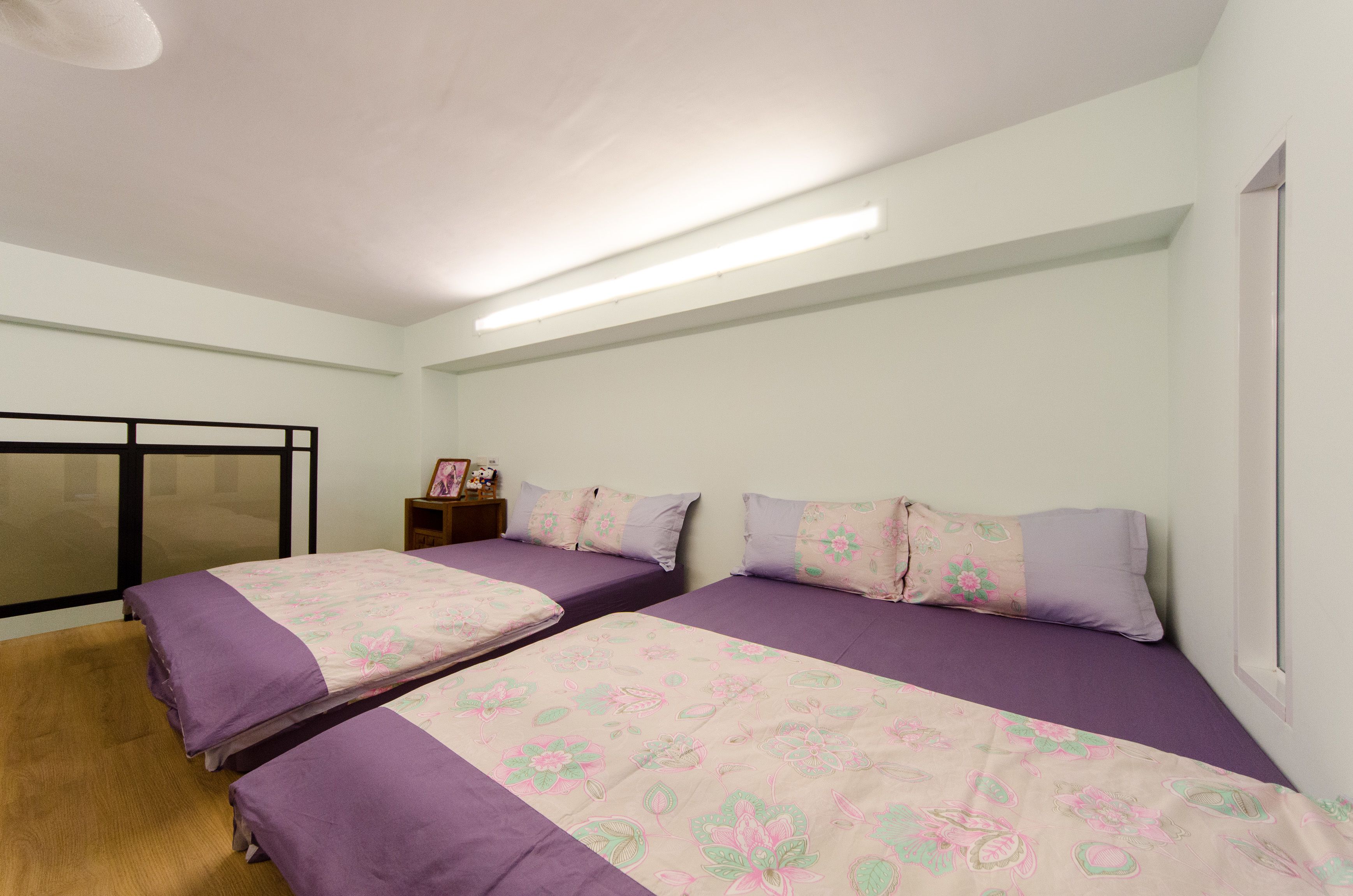 Habitación con dos camas, cada una con ropa de cama morada y motivos florales, suelo de madera, mesita de noche con lámpara y ventana con persianas.