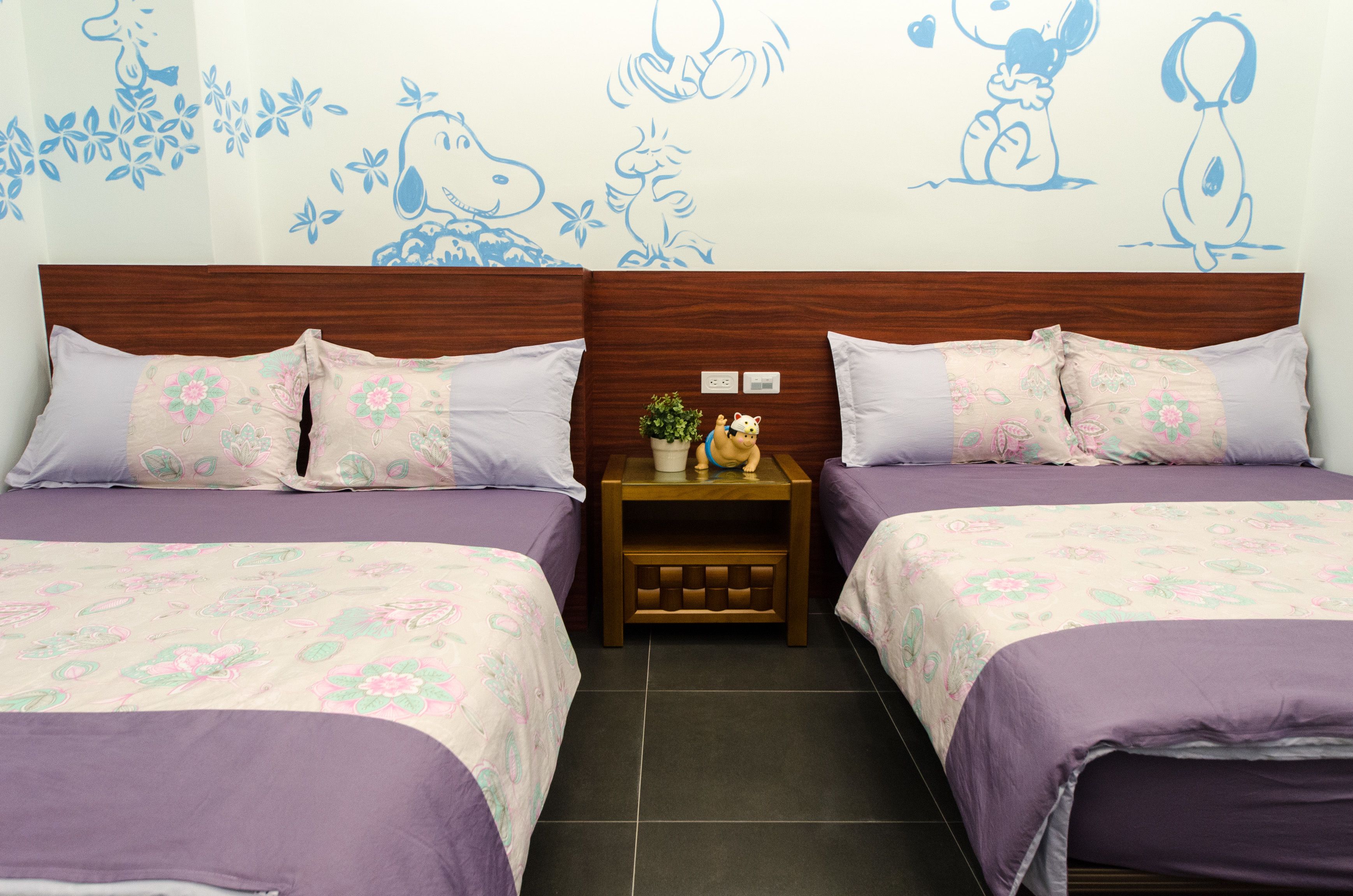 A hotel room with two beds, floral bedding, a nightstand with a plant and figurines, and wall art of cartoon characters.