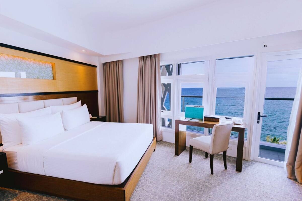 Ein Hotelzimmer mit einem großen Bett, einem Schreibtisch mit Stuhl und Meerblick aus den Fenstern.