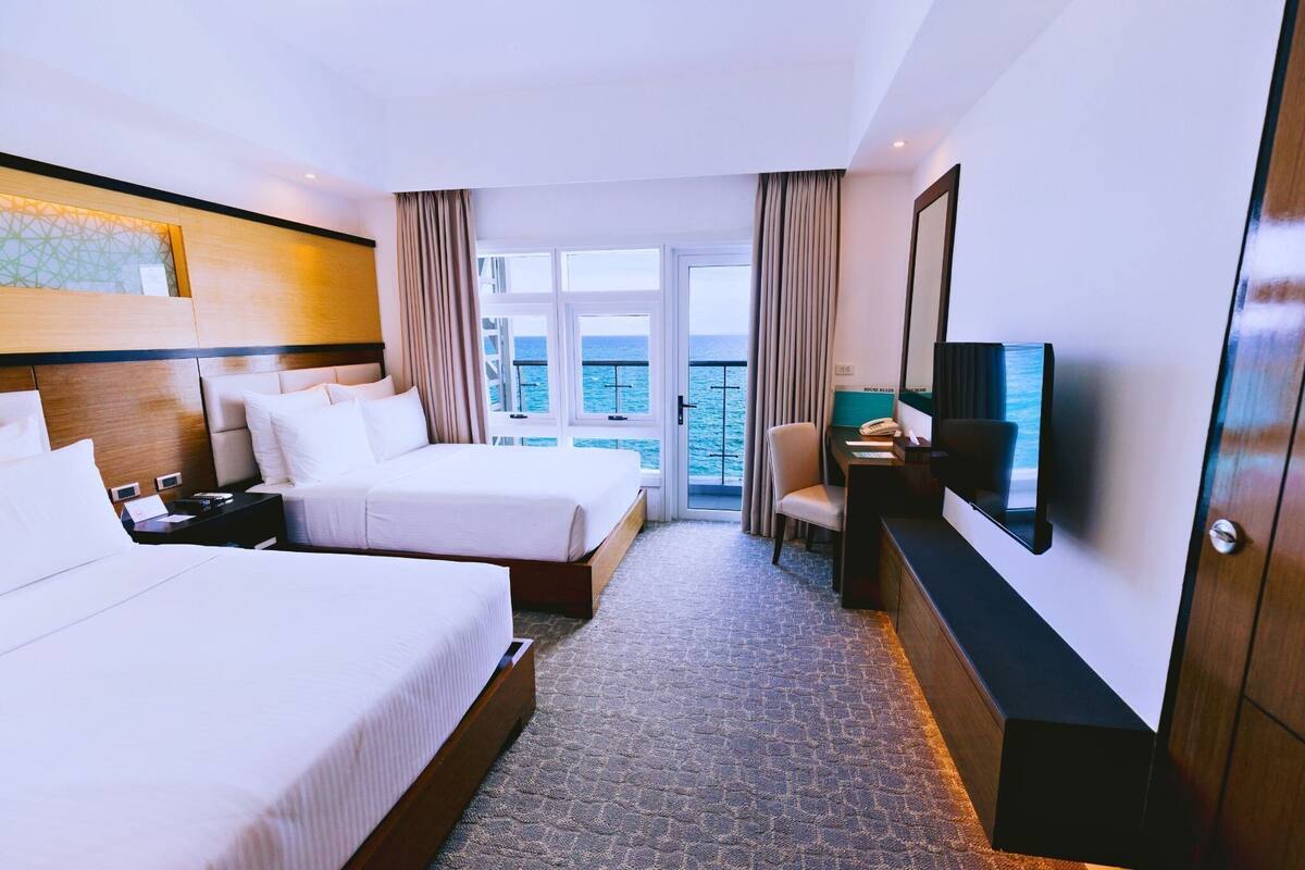 Ein Hotelzimmer mit zwei Betten, einem Fernseher, einem Schreibtisch und Meerblick.